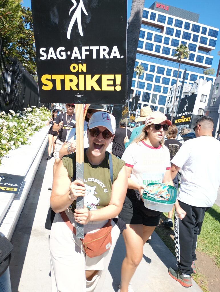 a woman in yellow in bright sunshine is smiling and holding a picket sign reading SAG AFTRA and WGA on strike!