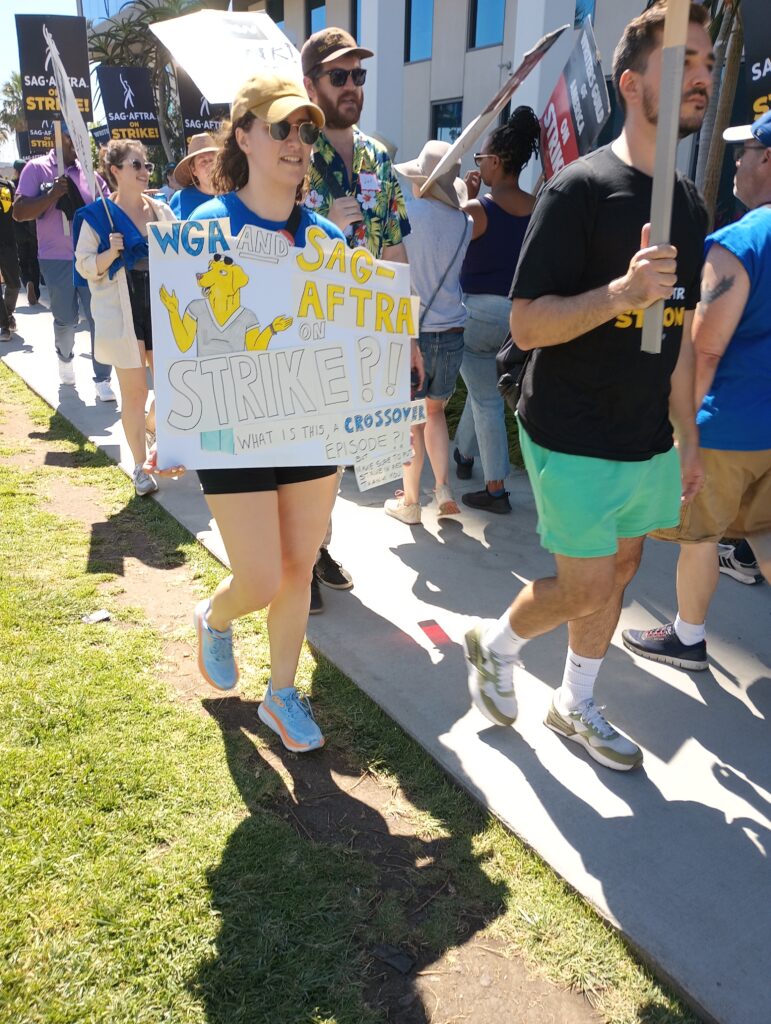 a picketer holds a white sign with yellow letters with the joke, what is this crossover episode? referring to unity between WGA and SAG-AFTRA.