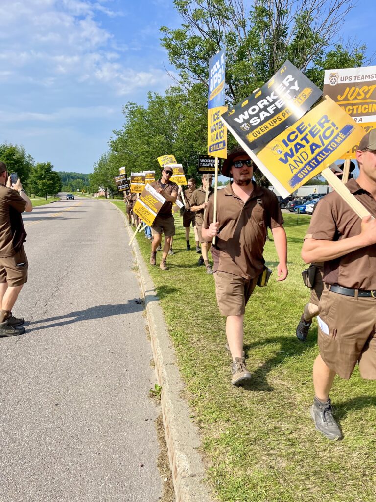 A line of about 25 UPS workers in brown shirts and shorts walks along a road carrying signs reading "Cool it, UPS! Ice Water and AC," "Worker Safety," and Just Practicing for a Just Contract