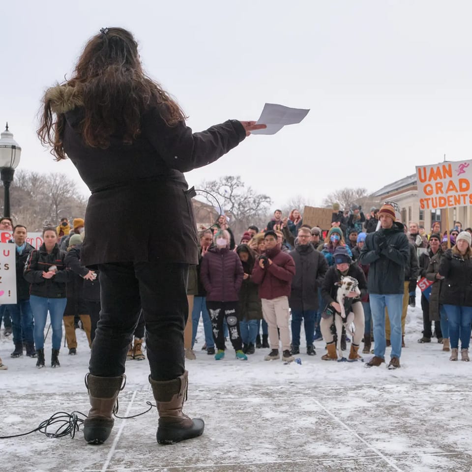 A person in a heavy coat stands speaking to others rallied around on a snowy day. The protesters hold signs identifying them as part of the University of Minnesota Graduate Labor Union.