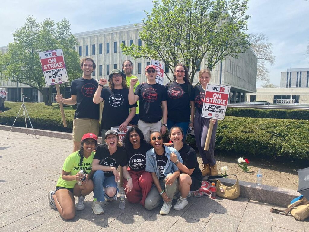A group of people kneel and stand in front of a university building holding picket signs and wearing shirts identifying them as Temple University Graduate Students Association.