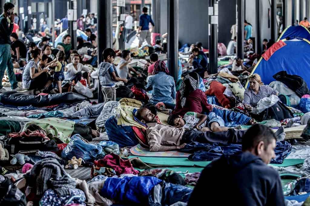 A large group of people seeking asylum at the U.S. border sit and lie down amid sleeping bags and other belongings. They look exhausted.