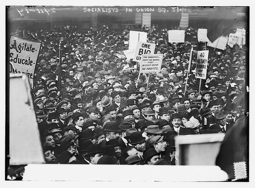 Thousands of people, mostly men in hats, are gathered in a public setting to listen to a speaker. They carry signs reading "Agitate, Educate, Organize," "One big union," and other slogans. The image is in black and white.