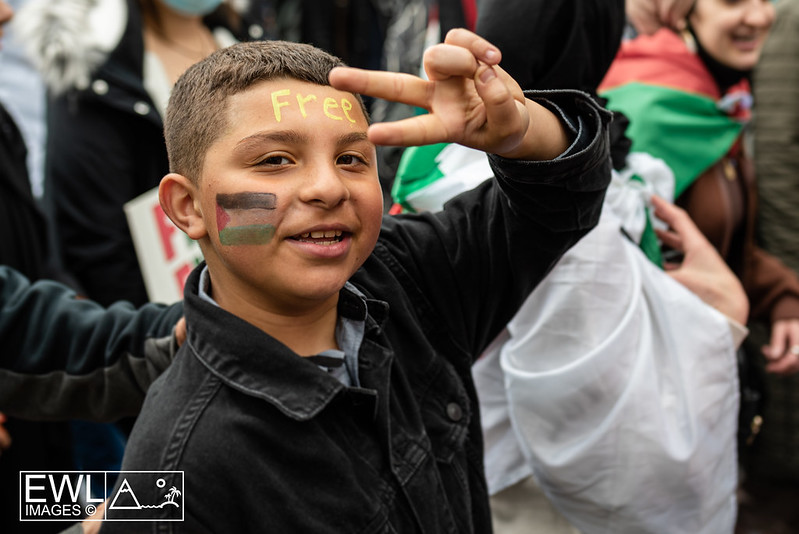 Boy in a London pro-Palestine protest in May 2021 during the Unity Intifada. His face is painted with a Palestine flag and the word “free” on his forehead. He is holding up his hands in the shape of a peace sign.