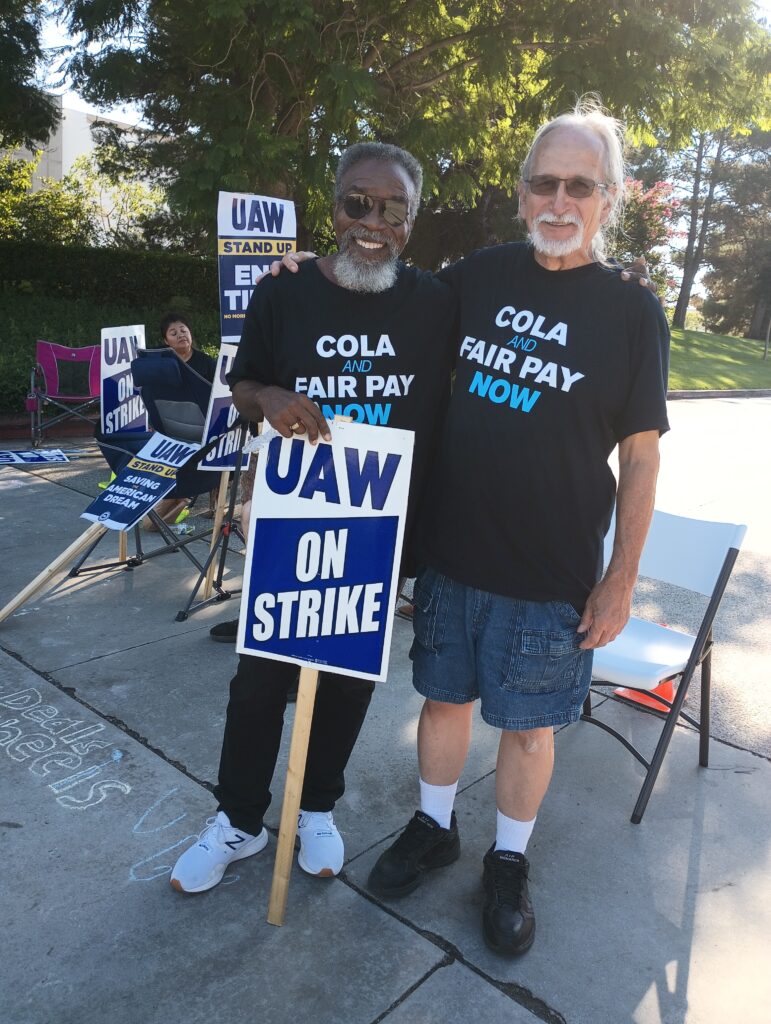 Two men, one Black in sunglasses and one white in sunglasses, stand in black t-shirts that read COLA AND FAIR PAY NOW. One holds a sign reading UAW ON STRIKE.