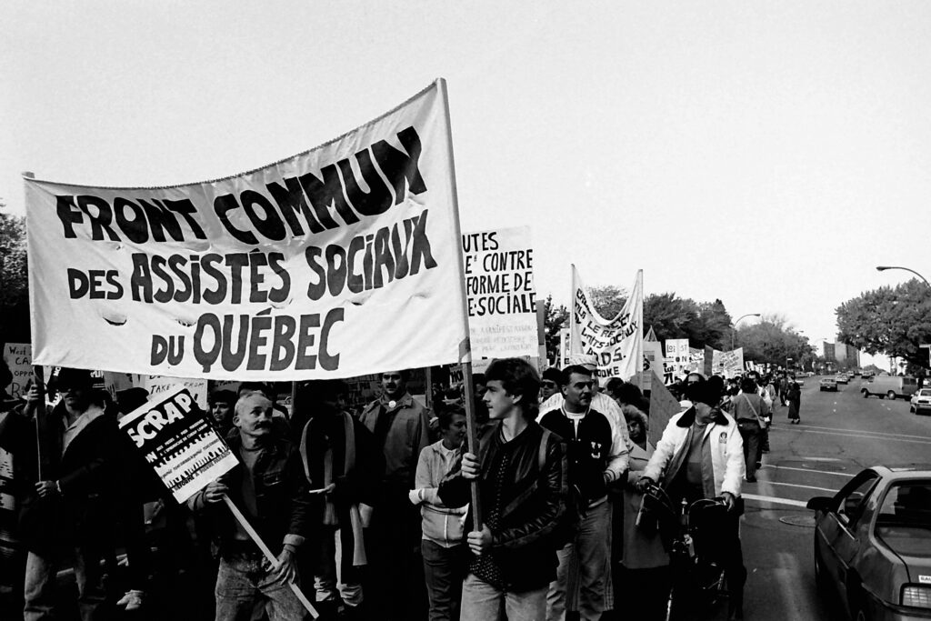 A black-and-white photo shows a line of marching demonstrators, stretching into the distance. Most are masculine-presenting and hoist protest signs and banners. Two workers in front hold a banner that reads, in French, “FRONT COMMUN DES ASSISTÉS SOCIAUX DU QUÉBEC.”
