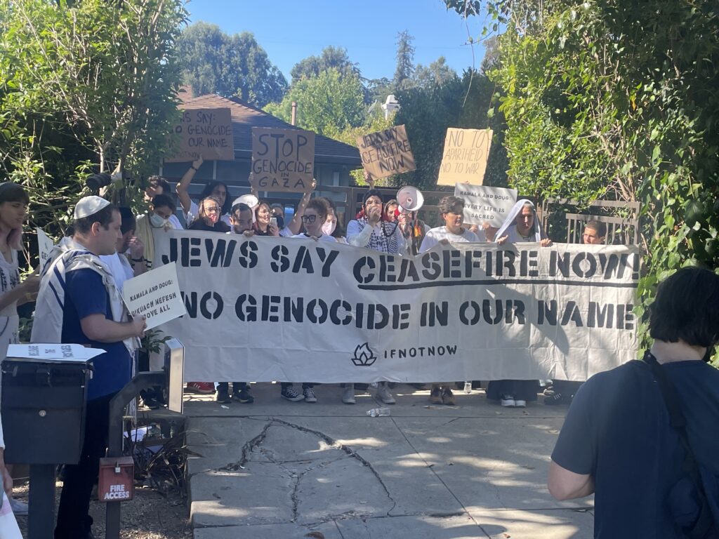 About fifty people holding signs reading Stop Genocide in Gaza stand behind a black-and-white banner reading Jews Say Ceasefire Now. No Genocide in our Name. IFNOT NOW