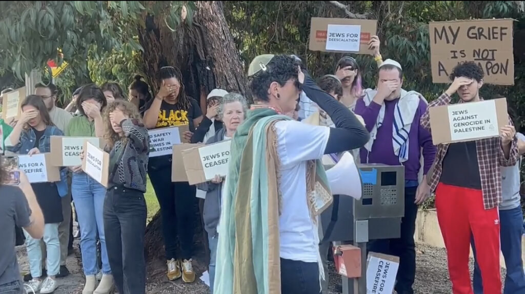 two dozen people carrying signs reading Jews for Ceasefire and "My Grief is not a Weapon" and wearing tallits and kippot, stand in prayer with their hands to their foreheads covering their eyes.