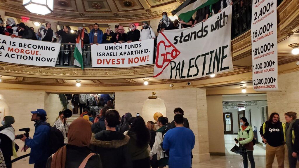 Large rally inside domed building atrium; banners reading GEO Stands With Palestine and Divest Now.