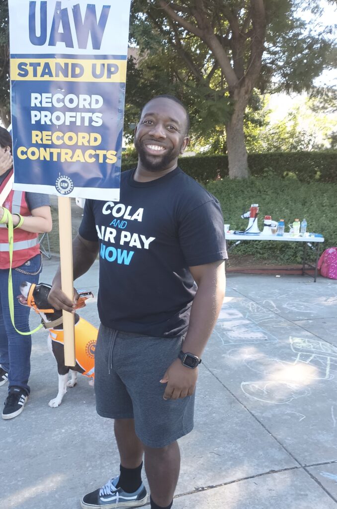 A black man holding a sign reading UAW STAND UP RECORD PROFITS RECORD CONTRACT and wearing a black t-shirt reading COLA END TIERS NOW 