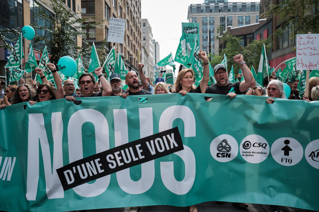 Workers, both feminine and masculine-pressenting, march behind a large green banner with their fists in the air. Behind them is a large crowd waving green flags. The banner reads, in French, “NOUS D’UNE SEULE VOIX” (“we are of one voice”) and features the names and logos of each union in the Front Commun (Common Front).