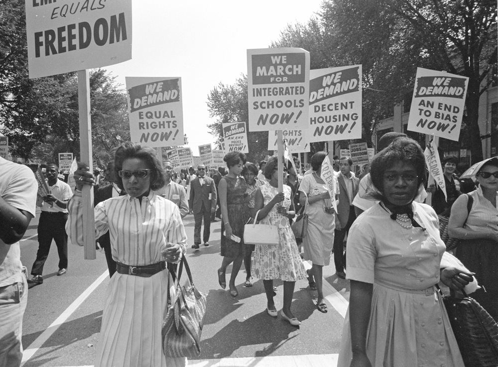Black and white photo. A predominantly Black crowd of women and men marches toward the camera down a tree-lined road. They carry printed signs with slogans such as, “We demand equal rights NOW,” “We march for integrated schools NOW,” “We demand an end to police brutality NOW,” and “We demand decent housing NOW.”