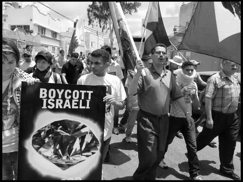 A protest in Bilin in the West Bank in 2010. A black and white photograph showing a march of protesters of different ages and genders carrying Palestinian flags and a poster calling for the Boycott of Israel and with a graphic of a large diamond. At this time, after the Friday noon prayers, villages throughout the West Bank dramatically and theatrically confront the occupation, including challenging the Israeli diamond trade, which accounts for one-third of Israel's export economy. Photo by scottmontreal
