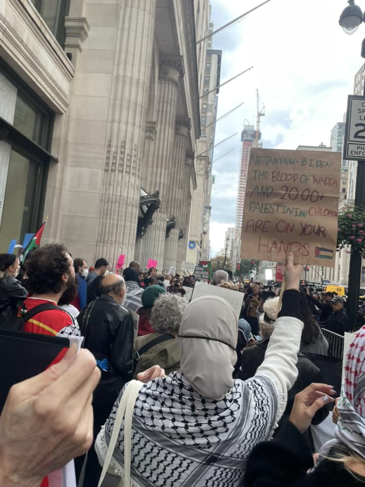 A crowd shot by behind marches down a NYC street. One carries a sign reading blood of Palestinian children on your hands.