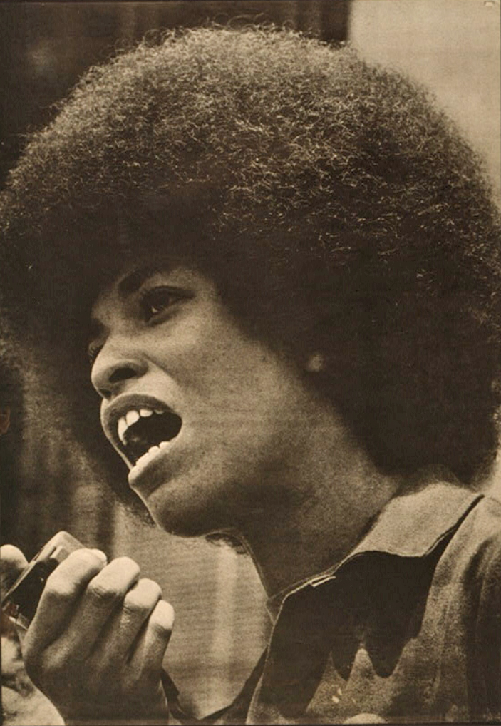 Sepia-toned black and white photo. A young Black woman with a large Afro haircut appears in a close-up three-quarter profile shot slightly from below. Her mouth is open as she speaks into a handheld microphone. The top-lighting suggests that she is outdoors, but the blurry background doesn’t give a clear indication.