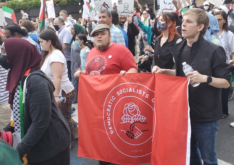Two members of the Charlotte Metro Chapter of the DSA marching at the March for Palestine in Downtown Charlotte, North Carolina, May 22, 2021, carrying a red DSA flag. Other protesters carrying pro-Palestine posters and flags. Photo by Bingjiefu He.