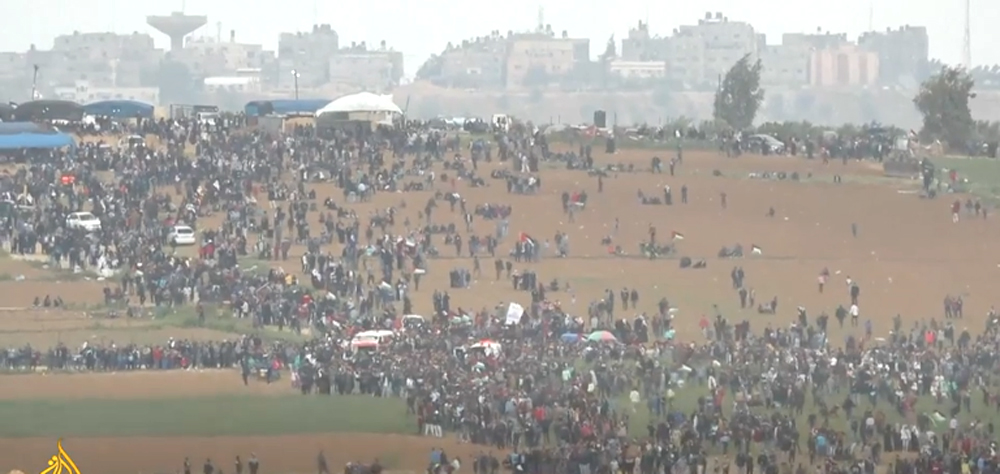 Long shot from behind a crowd of perhaps a thousand Palestinians in a open field approaching the fence confining Gaza. The day appears hazy. Beyond the fence are Israeli buildings of multiple stories; one of the taller ones looks like a watchtower.