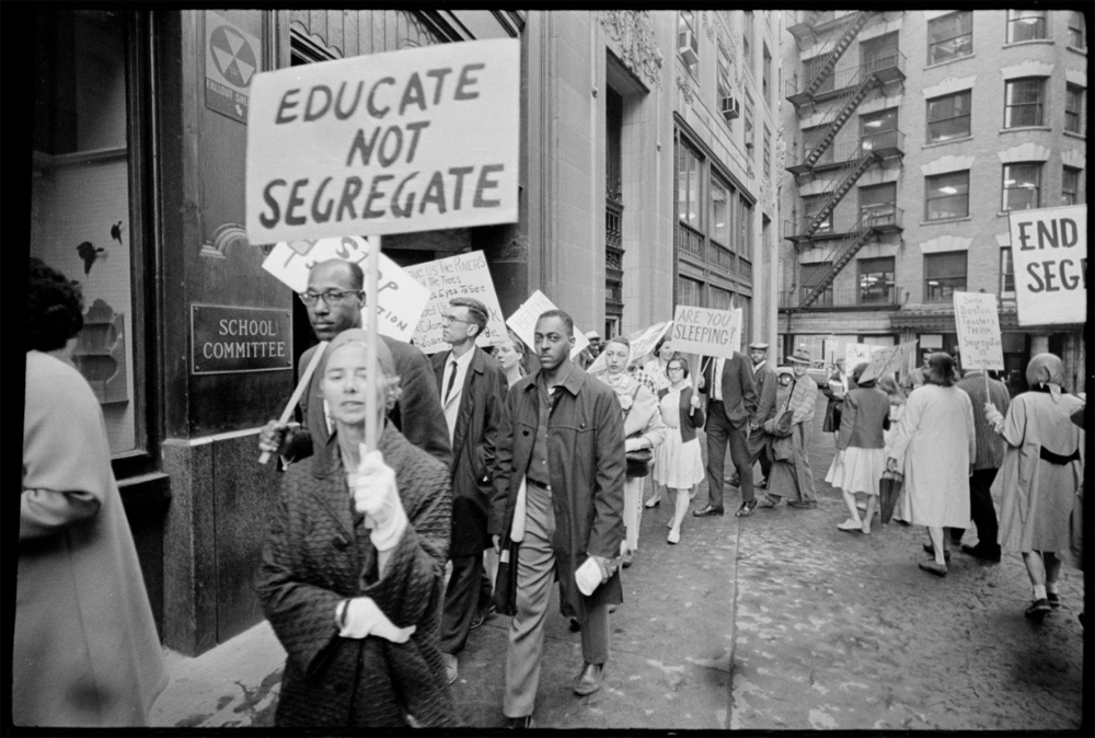 Black and white photo. Picketers march in a circle on the sidewalk in front of a building labeled, “School Committee.” The crowd is racially integrated and includes women and men. A woman in the foreground carries a hand-lettered sign that reads, “Educate not segregate,” while another sign in the background reads, “Are you sleeping?”