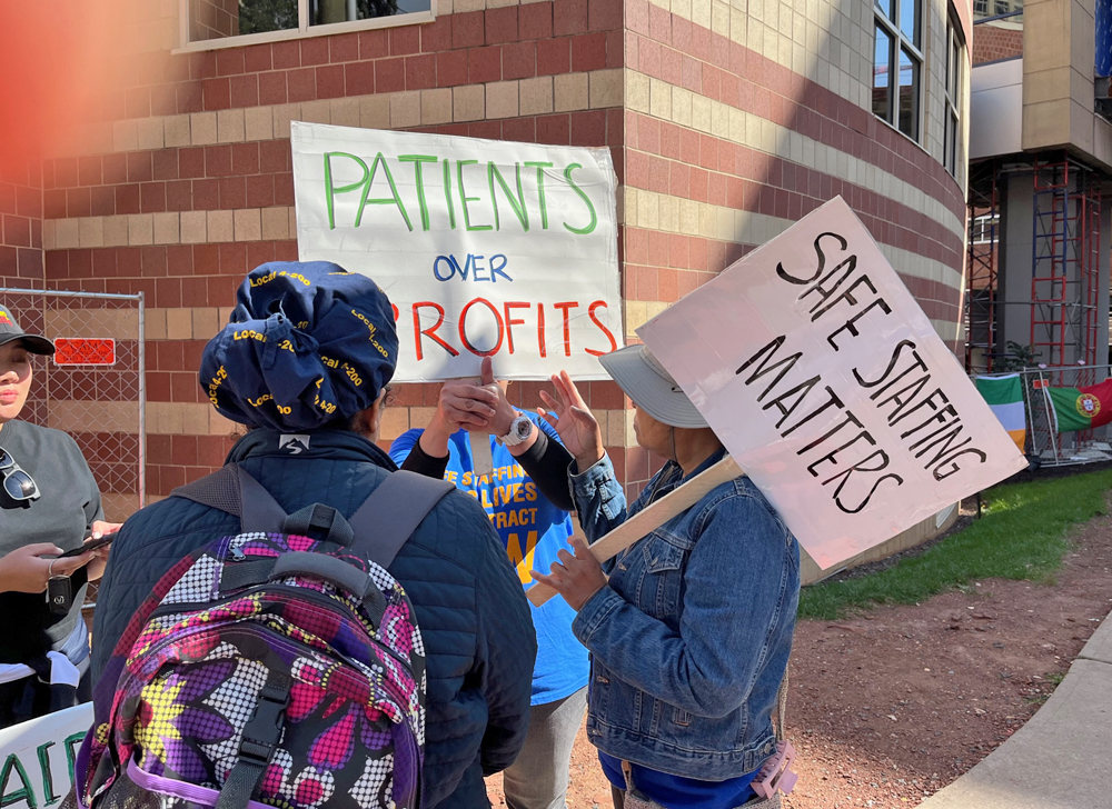 Picketers in font of a building hold signs reading Patients Over Profits and Safe Staffing Matters.