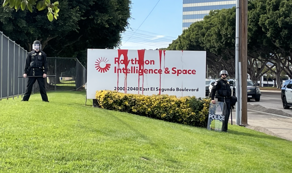 Two cops in riot gear flank the sign at the entrance of Raytheon Intelligence and Aerospace in El Segundo. There is red paint dripping from the white sign.
