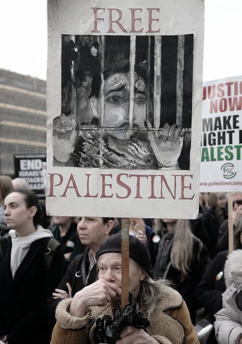 Artist and activist Lynda May Ayres holding her "Free Palestine" poster at an April 2018 Palestine demonstration in April 2018. The poster shows a the black and white image of a child behind prison bars with the word “Free” framing the top of the poster and the word “ Palestine” framing the bottom.
