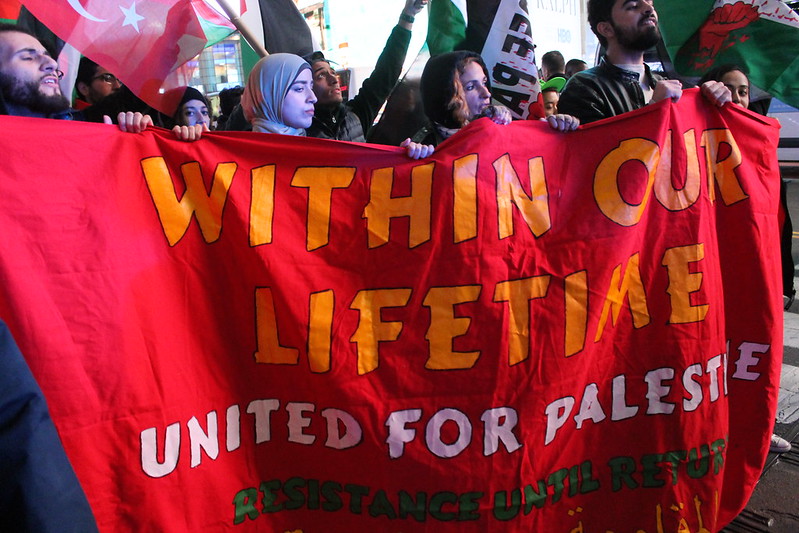 Red banner reading “Within our Lifetime”, “United for Palestine”, “Resistance Until Return”, from November 15, 2019 march in New York City to support Palestinians and resistance in Gaza. Photo by Joe Catron. The banner is held by four marchers with other banners in the background.