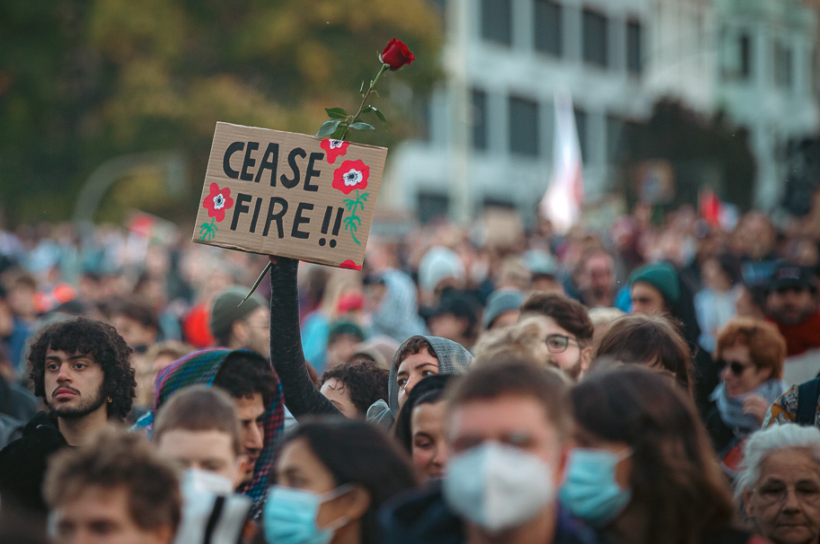 Photo of hundreds of demonstrators at a Free Palestine demonstration in Oranienplatz. Kreuzberg, Berlin, Germany, on October 21, 2023 . At the center of the photo is a protester holding a handmade cardboard sign reading, “ceasefire now” with two offset images of roses. The protester is also holding a rose with the sign.