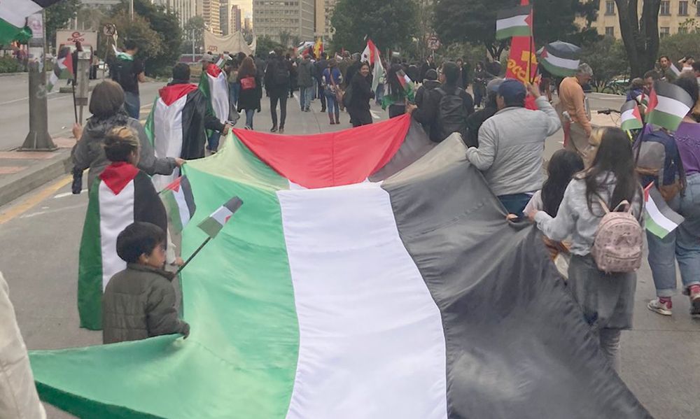 A long banner being carried horizontally in a street march, shot from behind. The banner, is green, white, red, and black—sewn together as a Palestinian flag. Several of the flag bearers are children, including young ones. Two people in the procession are draped in Palestinian flags, and other flags are carried on sticks.