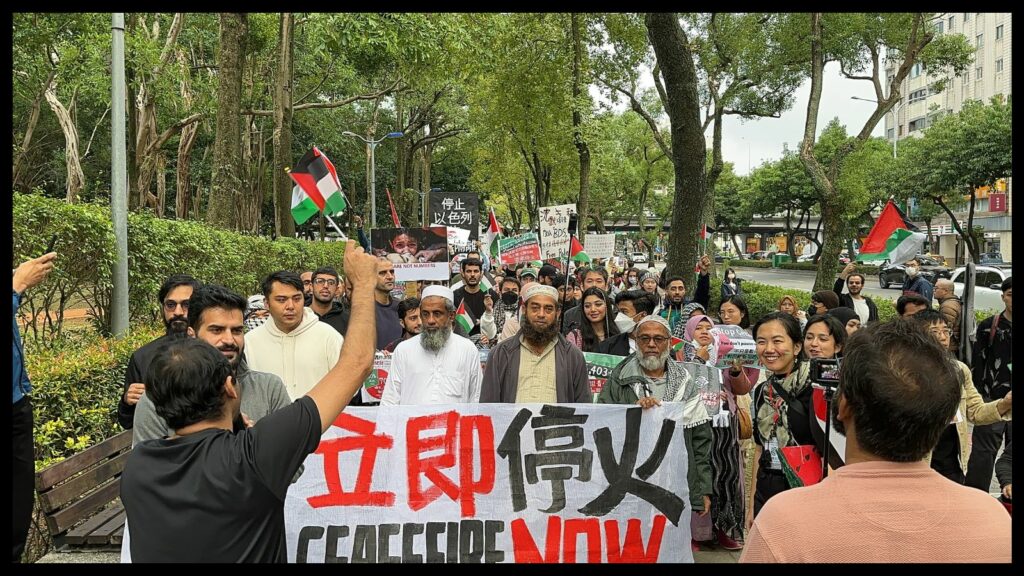 A line of protesters, many holding signs and waving Palestinian flags, stretches behind a banner with the words “Ceasefire Now” written in red and black in Mandarin and English. The crowd includes Muslim protesters who wear kufis and hijabs, and many protesters wear keffiyehs.