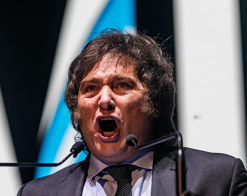 Photo of newly-elected Argentinian President Javier Milei with his trademark long hair and side-burns, mouth wide open as though screaming into two microphones framing his face.