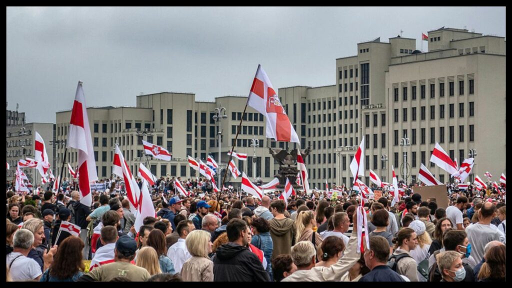 Under a gray sky, a massive crowd demonstrates in front of sprawling gray building. Most of the crowd is facing away from the camera, toward the building. People are holding multiple, large, red and white Belarusian flags that wave left to right above the crowd.