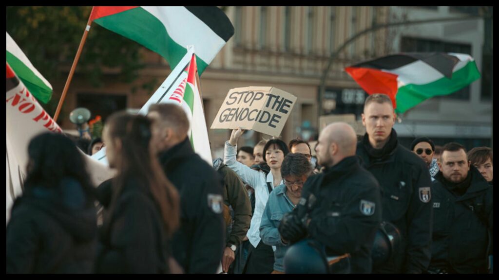 Color photo of demonstrators on a city street. Several Palestinian flags are waving in breeze, which is blowing from left to right. The foreground is dark and slightly blurred, with a few cops on the right. One woman—a bit farther away at center of the photo—is in sharp focus wearing a light-colored shirt. She is framed by the darker elements, and she holds up a hand-lettered sign that reads, “STOP THE GENOCIDE.”