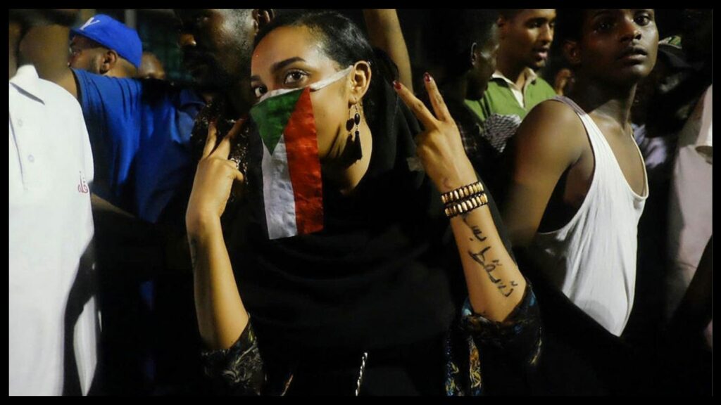 Close nighttime shot of Sudanese demonstrators. A woman in the center wears a small Sudanese flag—red, green, white, and black—as a veil covering her mouth and nose. She holds up her forearms, showing a the words “Just fall” in Arabic on her left arm.