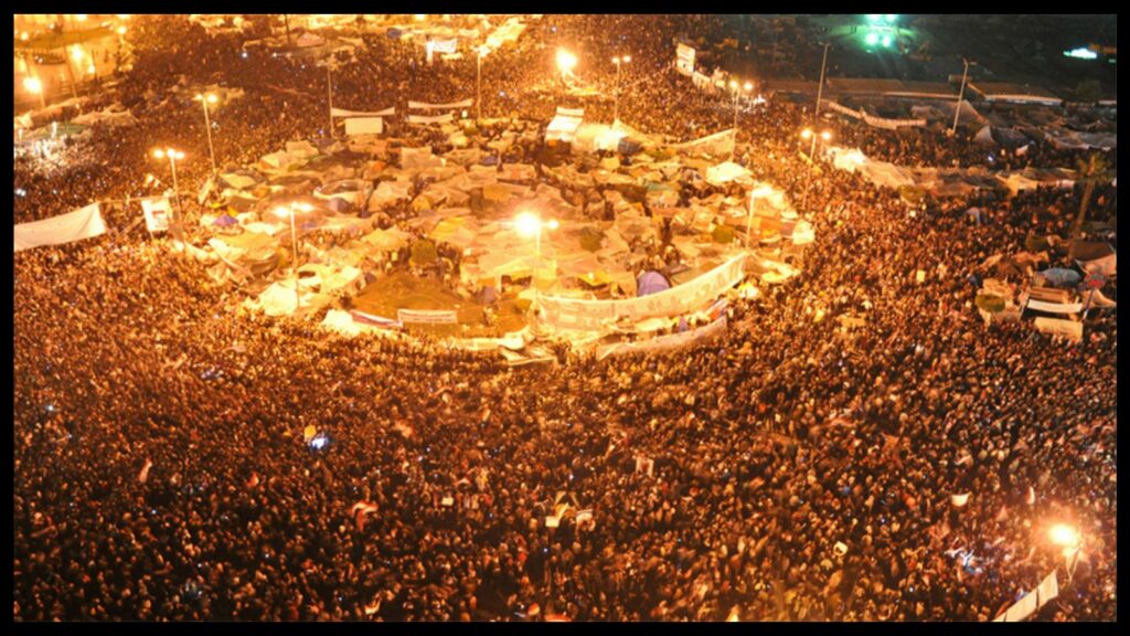 Nighttime aerial view of a large public square with a circular tent city in the middle. A crowd of thousands is packed around the tents. The crowd extends outside the frame of the picture in all directions and is illuminated by bright yellow light.