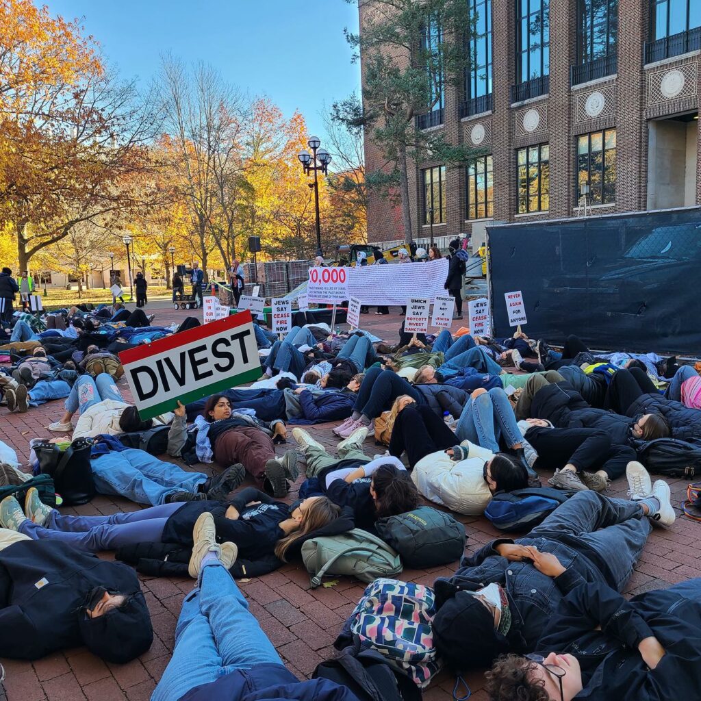 about 75 people lie on a campus plaza under fall trees as if dead. Some hold signs reading "Divest."