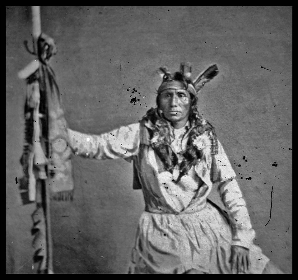A black and white photograph of a Dakota tribal chief Little Crow in a feathered headdress holding onto a staff.