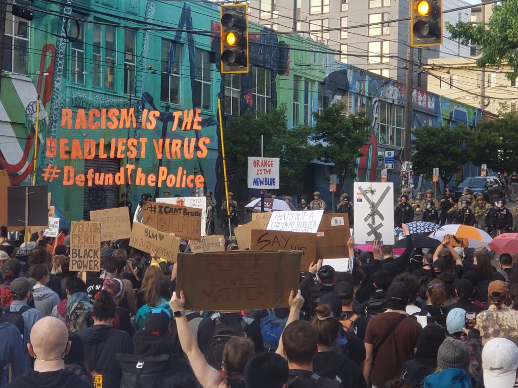 A large banner that reads “Racism is the deadliest virus. #Defundthepolice” is held aloft above a large crowd marching in the street toward a stoplight intersection.