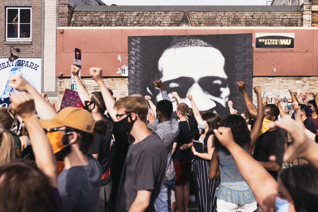 A multiracial crowd raises its fists outside a brick building, the side of which features a massive black and white portrait of George Floyd.