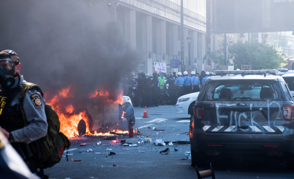A photo depicts a police car ablaze with flames and heavy smoke billowing from its roof. To the left, a uniformed cop walks away from the scene wearing a gas mask; behind the car is a crowd of protesters facing a line of police, with one protester holding a sign that reads “F**K THEM KKKOPS;” to the right, the trunk of a police van has been spraypainted with the words “FUCK 12.”