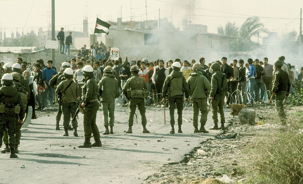 Color photo on a rubble-strewn road with weeds off to the right. The shot is taken from behind about a dozen soldiers, many with white helmets. They carry truncheons and assault rifles. Facing them across a razor-wire barrier is a crowd of fifty or more unarmed young people, apparently all boys. Smoke rises just behind the crowd. One protester holds up a picture that appears to be of Yasser Arafat; another holds up a Palestinian flag. Some distance back, two boys stand on a the wall of a crude one-story structure. One of them has a posture as if he is following through on throwing a rock toward the soldiers.