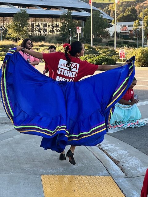 A brown-skinned dancer, photographed from behind, leaps from the pavement with her blue skirt spread wide like wings. She is wearing a shirt reading CFA Strike Ready! Campus buildings are in the background.