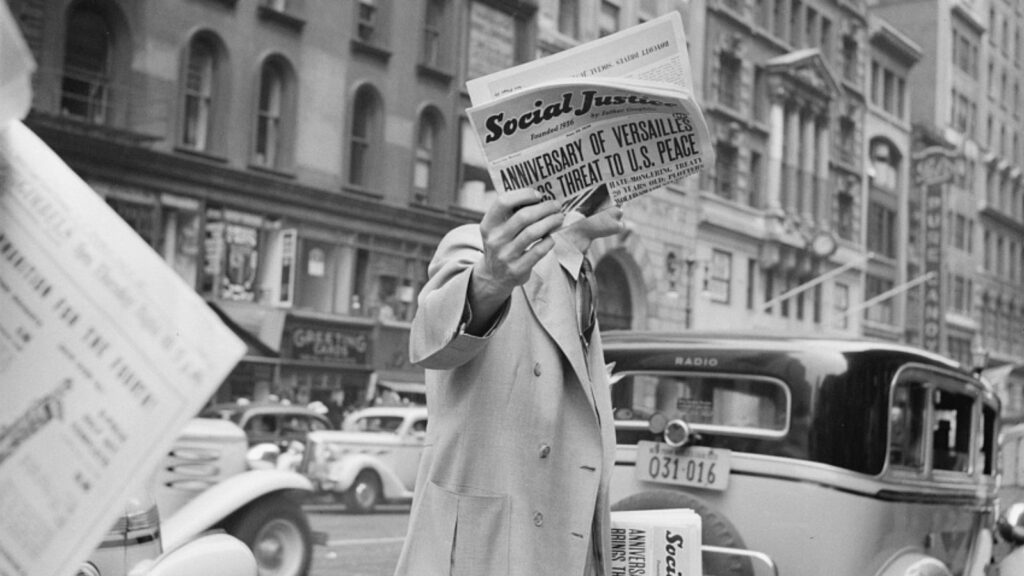 A man is displaying a copy of the newspaper Social Justice founded by Father Coughlin in his right hand while holding additional copies of the newspaper in his left hand. He is standing by two vehicles in front of a busy New York City street in the 1930s.