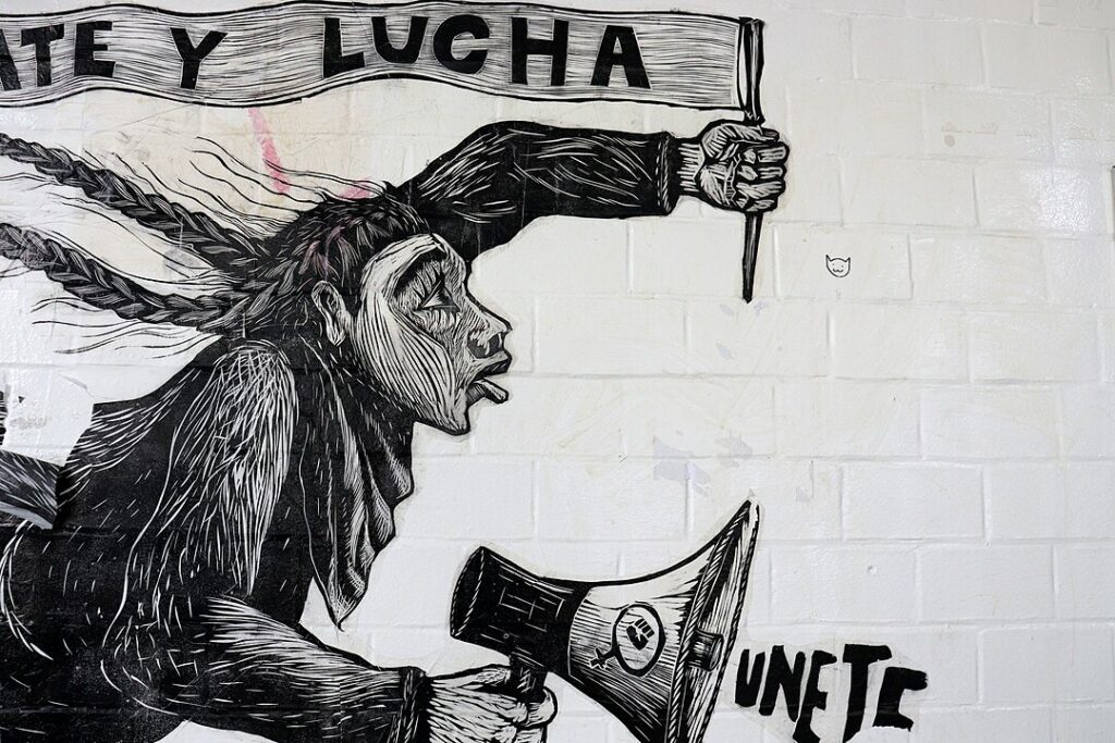 Photo of part of a mural from the Facultad de Filosofía y Letras de la UNAM. The image in the mural shows a student, in motion moving left, carrying a banner in one hand and a loudspeaker in the other hand. The mural shows the partial slogan ending in “struggle” (in Spanish, “lucha”) and the loudspeaker has a fist logo on it with writing which reads: “join” (in Spanish, “unete”).