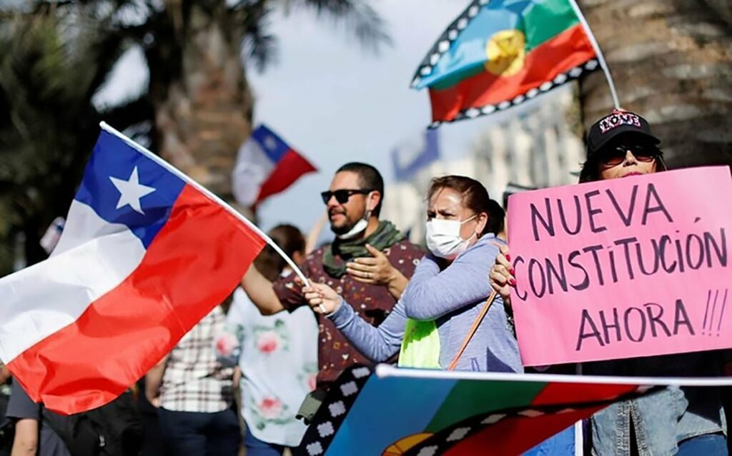A small crowd is gathered outside in support of a new political constitution for the country of Chile. Three individuals are featured in the forefront, a man wearing a multicolored purplish shirt, a white surgical mask hangs around his neck along with a green scarf and he is gesturing positively with his hands, next to his left is a woman wearing a white surgical mask, blush shirt is waving a Chilean flag with her right hand while her left arm is crossed over her right shoulder, and another woman is on her left wearing a black hat that reads love in colorful lettering, a pair of sunglasses, jeans, and is holding a pink sign that reads in all caps “NUEVA CONSTITUCION AHORA” that translate to New Consituiton Now”
