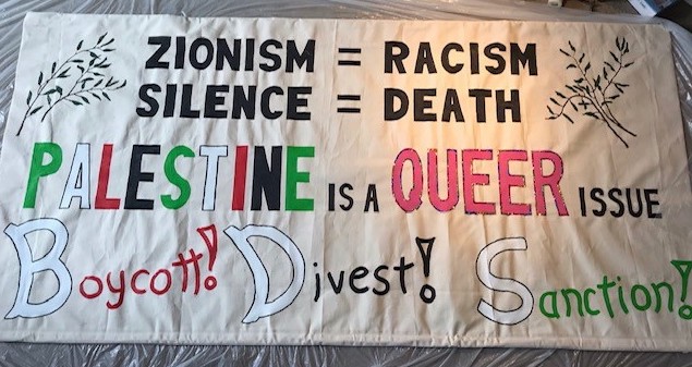 Banner of NYC Queers Against Israeli Apartheid,  circa 2019, which reads, in black lettering at the top, “Zionism equals racism” and just below, also in the same black lettering, “Silence equals death.” Across the middle of the banner it reads: “Palestine is a queer issue.” The word “Palestine” is written in alternating  white, green, red,  and black of the Palestinian flag. The word “queer” is written in pink lettering. The bottom of the banner reads: “Boycott,” “Divest,” “Sanction” with each initial letter in white, with the rest of the first word in red, the rest of the second word in black, and the rest of the third word in green. Images of olive branches decorate the upper left and right corners of the banner.
