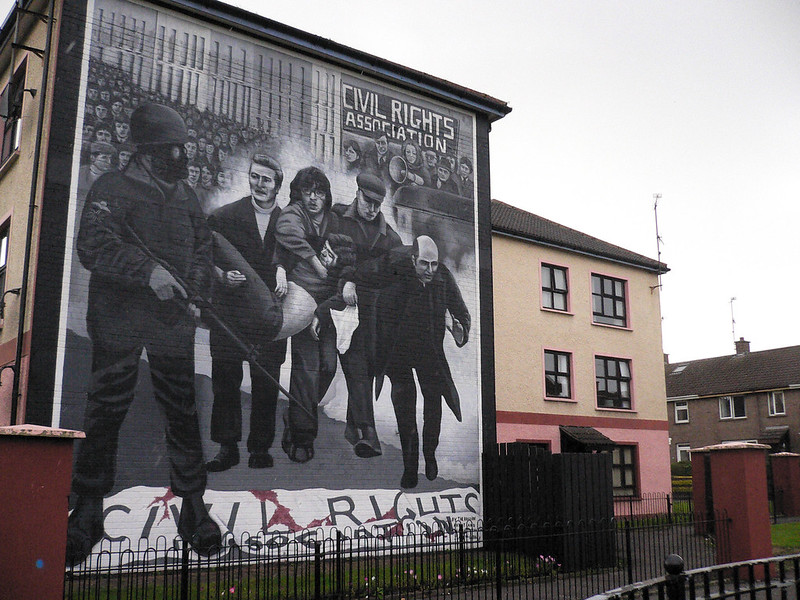 Large square outdoor mural on a brick building. The image is mostly black-and-white, showing civilians carrying a bloody and wounded comrade. A figure with an automatic rifle, a gas mask, and an arm patch of the Union Jack, looks on. This soldier is trampling a bloodied banner that reads, “CIVIL RIGHTS ASSOCIATION.”