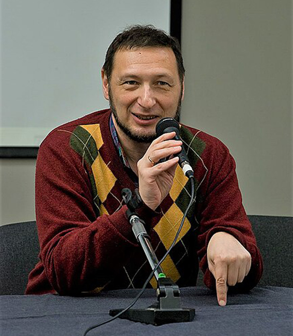 Indoor torso-and-head shot of a man speaking into a microphone. He has a beard with no mustache and is wearing a pullover sweater.