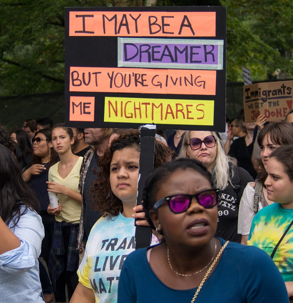 A crowd of protesters, mostly women. In the foreground, a Black woman in a navy shirt and sunglasses. Behind her, a serious looking woman with curly brown hair holds a multi-colored sign reading I may be a dreamer, but you're giving me nightmares.