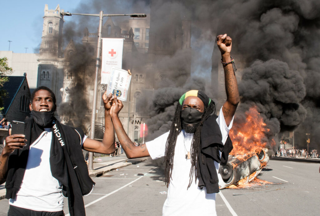 Two Black people clasp hands as they hold up a Philadelphia police armband in front of a burning police cruiser. They wear white tshirts and Black protective masks. There is a church in the background behind billowing smoke.