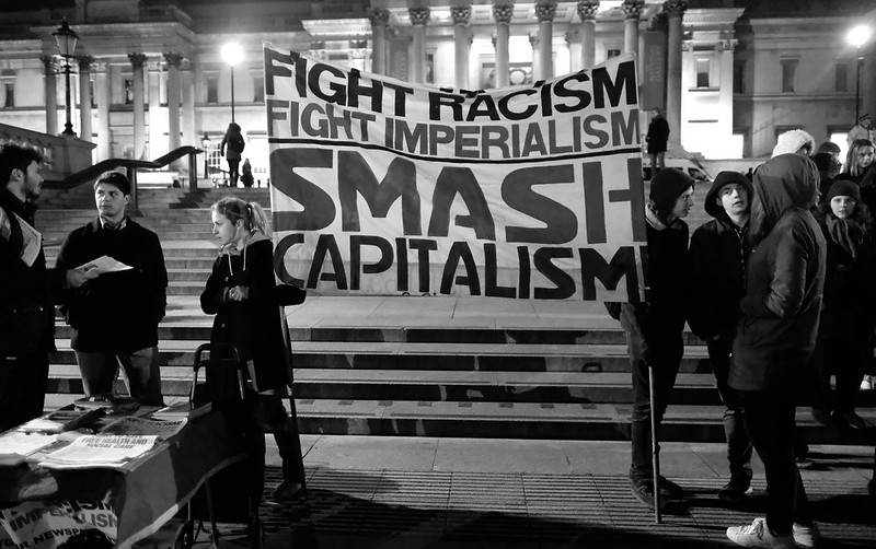 A black and white photo of about 8 activists at night in a lit-up London's Trafalgar Square. They hold a banner that reads in capital letters Fight racism, fight imperialism, smash capitalism. There is a literature table to the left where people are talking about a leaflet.